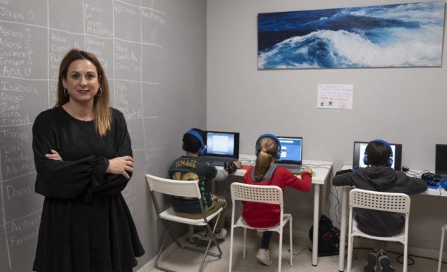 Una mujer está de pie con los brazos cruzados junto a un grupo de niños que utilizan el método WITS mientras trabajan en las computadoras. En la pared de la izquierda hay una pizarra con escritura y un cuadro de olas cuelga de la pared del fondo.