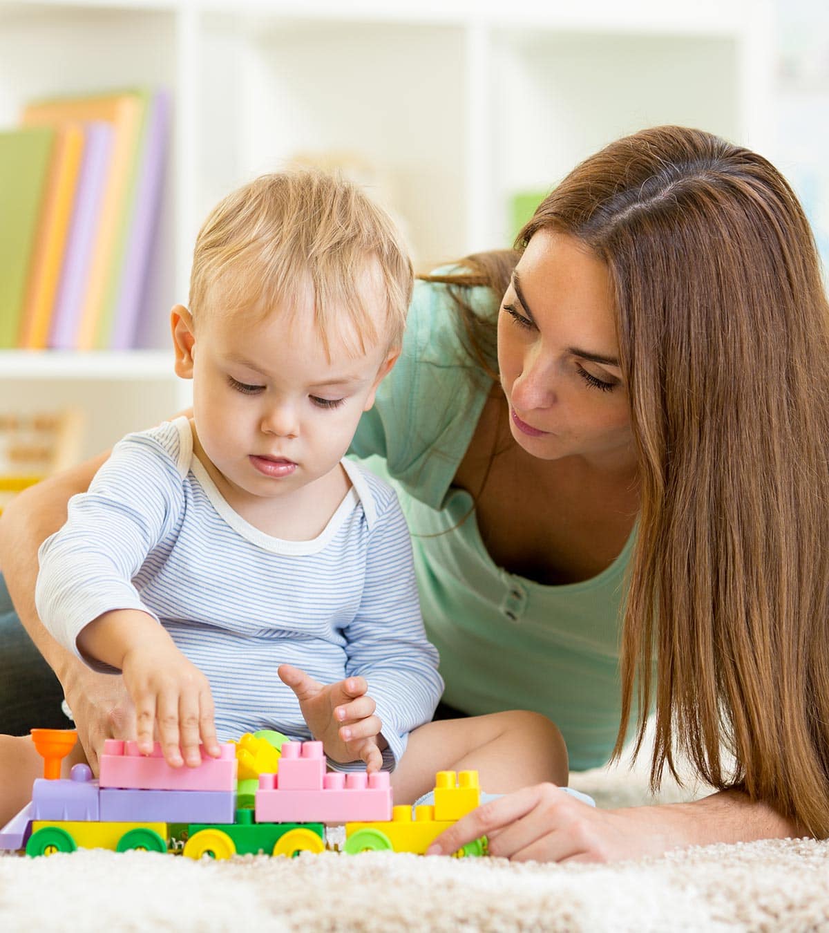 Una mujer y un niño de entre 18 y 24 meses están sentados en el suelo, jugando con bloques de plástico de colores con forma de tren. Al fondo se sitúan estanterías con libros y juguetes, creando un ambiente acogedor durante el confinamiento.