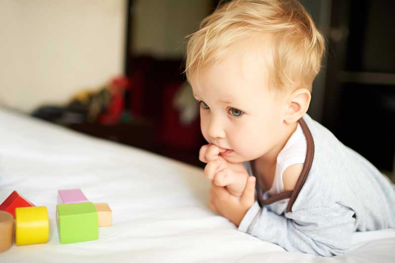 Un niño pequeño de cabello rubio y camisa gris yace sobre una cama blanca, mirando fijamente los coloridos bloques de madera, típicos de LA EDAD DE LOS DOS AÑOS, colocados frente a él.