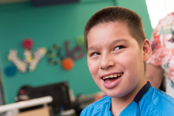 Un niño sonriente con una camisa azul, con el fondo borroso del aula y decoraciones coloridas en las paredes, representa la alegría de aprender a pesar de vivir con EL SÍNDROME DE ANGELMAN.