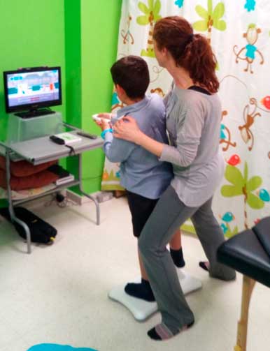 Una mujer ayuda a un niño a usar una consola de juegos Wii en una habitación luminosa y colorida con paredes verdes y cortinas con temas de monos. El niño se para sobre una tabla de equilibrio blanco mientras sostiene un control remoto, integrando la diversión en su sesión de fisioterapia.