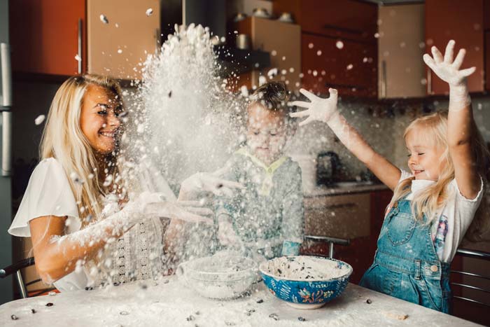 Tres hijos se divierten en una cocina durante el verano, jugando con la harina que se lanza al aire, creando una nube de polvo blanco a su alrededor. Estos momentos lúdicos también cuentan como actividades beneficiosas, fomentando la creatividad y el vínculo.