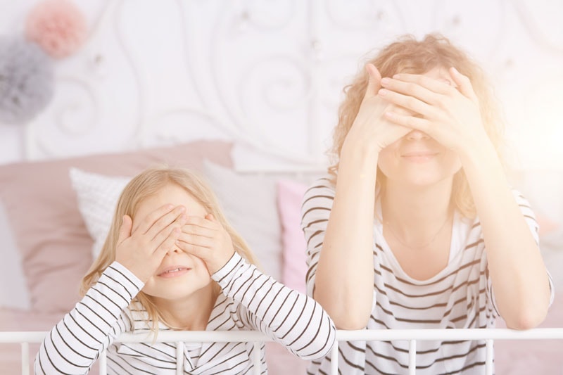 Una mujer y una niña, ambas con camisas a rayas, se cubren los ojos con las manos mientras están sentadas en una cama. Como dice el refrán, "LOS NIÑOS VEN, LOS NIÑOS HACEN.