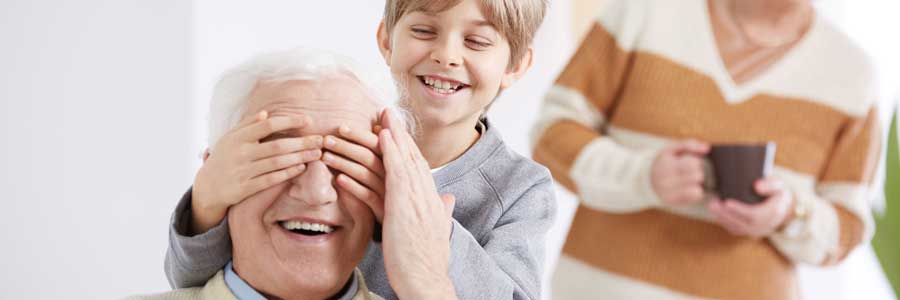 Un niño tapa los ojos de una persona mayor que sonríe celebrando el Día de los Abuelos, mientras otro adulto sostiene una taza al fondo.