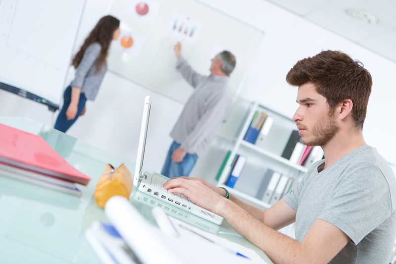 Un adolescente está sentado en un escritorio trabajando en una computadora portátil. Al fondo, dos personas, un hombre y una mujer, están paradas cerca de una pizarra con gráficos que analizan las opciones de tratamiento para el tartamudez.