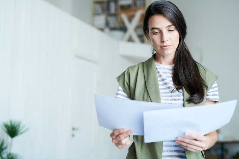 Una mujer se encuentra en una habitación bien iluminada, sosteniendo y leyendo dos hojas de papel. Tiene el pelo largo y oscuro y lleva un chaleco verde sobre una camisa a rayas. Como adulta con dislexia, está revisando materiales para comprender mejor las estrategias efectivas para controlar su afección.