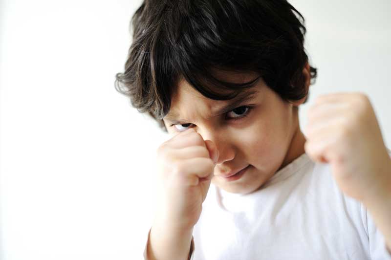 Un niño de cabello oscuro y expresión seria levanta ambos puños como si estuviera listo para boxear, vestido con una camiseta blanca, sobre un fondo liso y blanco. Esta imagen evoca preguntas sobre conductas agresivas y cómo actuar efectivamente como padres de hijos que exhiben tal comportamiento.