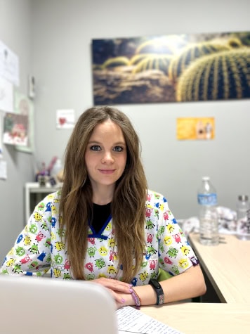 Una mujer con el pelo largo y una bata colorida se sienta en un escritorio en una oficina. El fondo presenta obras de arte con temática de cactus y una botella de agua descansa sobre el escritorio. El entorno tranquilo refleja los valores de comodidad y estilo de Nosotras en los entornos de trabajo cotidianos.