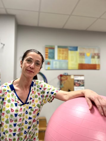 Una mujer con una colorida camiseta de Nosotras se encuentra junto a una pelota de ejercicio rosa en una habitación con un techo de azulejos y obras de arte abstractas en la pared detrás de ella.