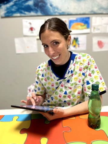 Una mujer que lleva una blusa médica colorida sonríe y está sentada en una mesa con una tableta y una botella verde. Nosotras puede ver dibujos infantiles en la pared detrás de ella.