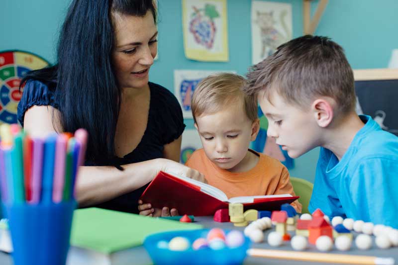 Un adulto lee un libro a dos niños pequeños en un salón de clases, con coloridos materiales educativos en la mesa a su alrededor y obras de arte expuestas al fondo. La sesión tiene como objetivo apoyar a aquellos con problemas de aprendizaje, creando un ambiente inclusivo y atractivo para todos los estudiantes.