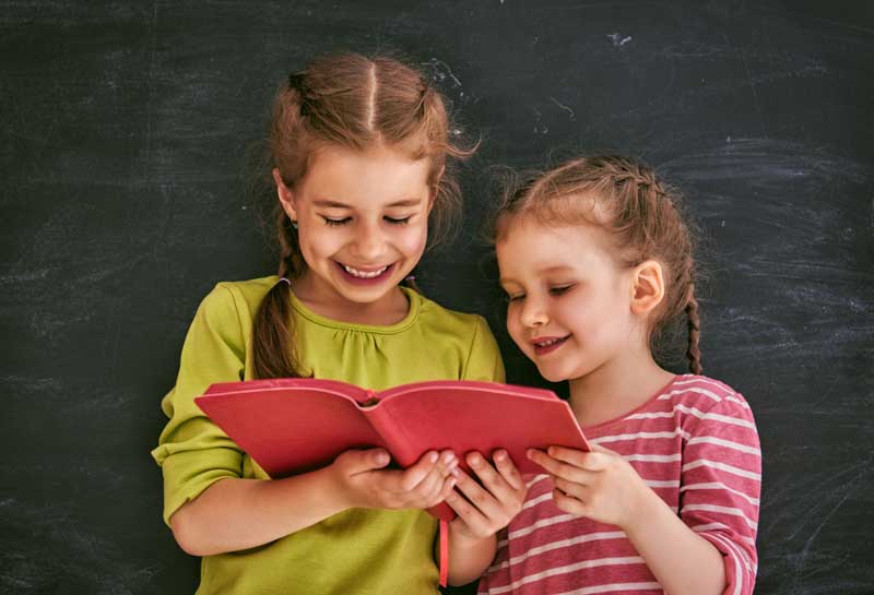 Dos jóvenes de pelo trenzado sonríen mientras leen juntas un libro rojo, superando cualquier dificultad de aprendizaje. Detrás de ellos se ve un fondo de pizarra.