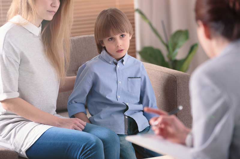 Un niño se sienta junto a una mujer, posiblemente uno de sus padres, durante una reunión con un profesional en una oficina. La mujer tiene su mano sobre el brazo del niño mientras el profesional toma notas y analiza pautas educativas para apoyar el aprendizaje del alumno.