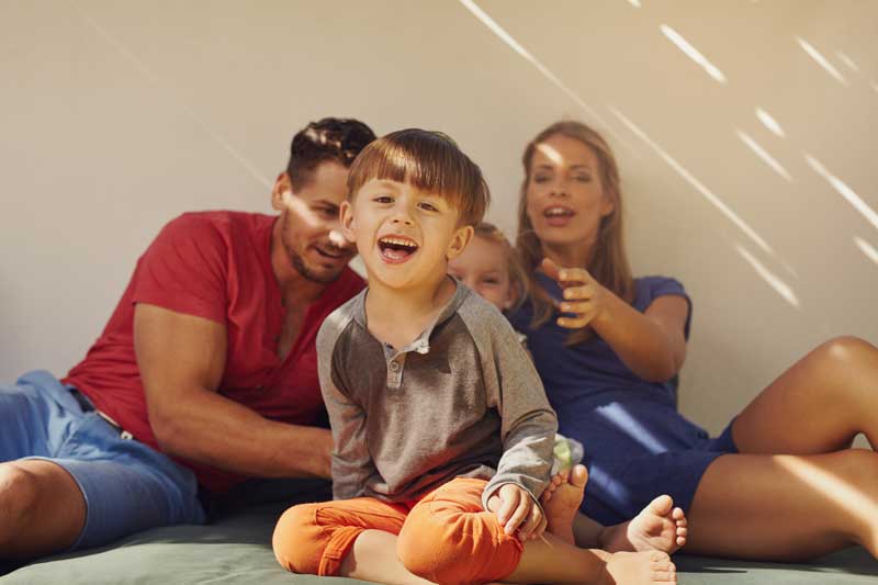 Una familia sentada en el suelo, sonriendo. Un niño delante se ríe, y detrás un hombre y una mujer, ambos también sonriendo. Se ve parcialmente el rostro de otro niño, contribuyendo alegremente a sus entornos competentes.