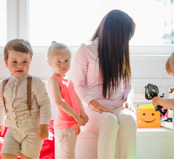 Tres niños y un adulto se encuentran en una habitación luminosa. En primer plano hay dos niños, uno con tirantes y el otro con un top rosa. El adulto está sentado, probablemente ofreciendo orientación similar a la psicología en Sevilla, mientras otro niño juega con un juguete cerca.