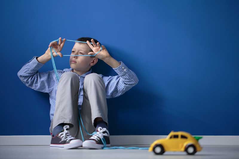 Un niño está sentado en el suelo, contra una pared azul, jugando con una cuerda. Frente a él hay un pequeño coche de juguete amarillo en el suelo, que podría ser uno de los principales signos de los trastornos del espectro autista.