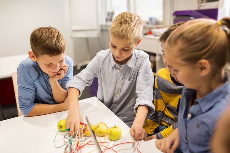 Cuatro niños se reúnen alrededor de una mesa con un proyecto de ciencias que involucra cables conectados a limones, fomentando la autoestima mientras colaboran y fortalecen su amistad.