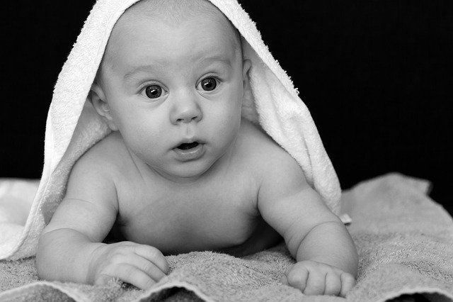 Un bebé con los ojos muy abiertos se acuesta sobre una toalla, parcialmente cubierto con otra toalla sobre su cabeza. La imagen, en blanco y negro, captura maravillosamente la esencia del desarrollo infantil durante los primeros años.