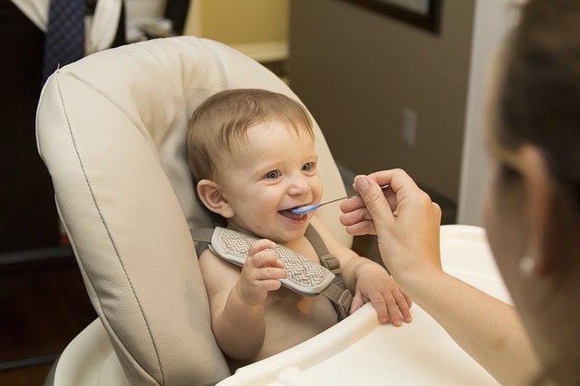 Un bebé sonriendo mientras lo alimentan con cuchara, sentado en una silla alta, y mirando a una persona, disfrutando del momento y beneficiándose de una nutrición adecuada.