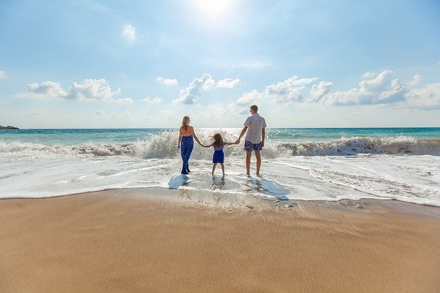 Una familia de tres personas se toma de la mano mientras está de pie al borde de una playa de arena, frente al océano con olas rompiendo suavemente, capturando la esencia del verano y la pura felicidad.