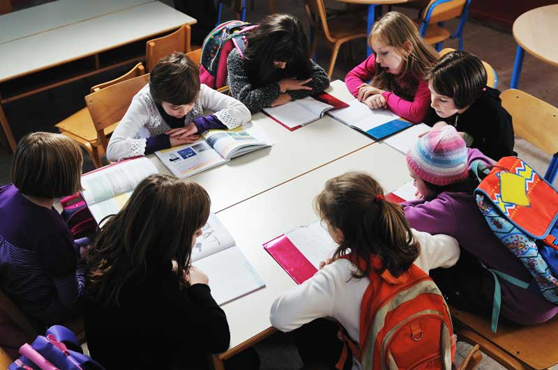 Un grupo de niños se sientan alrededor de una mesa en un salón de clases, leen libros y estudian juntos. Las mochilas son visibles en sus sillas y el salón de clases tiene un aire de dedicación mientras trabajan duro para lograr sus objetivos. Algunos incluso pueden ser beneficiarios de la Beca de Necesidades Educativas Especiales 2018/2019.