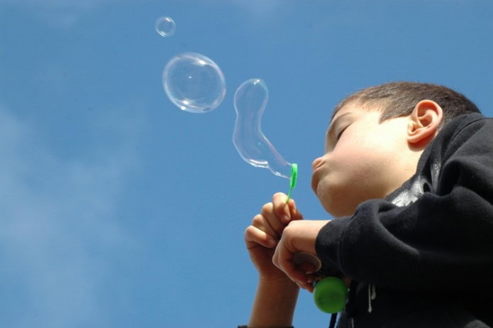 Un niño participa alegremente en ejercicios de soplo, soplando pompas de jabón bajo un cielo azul despejado.