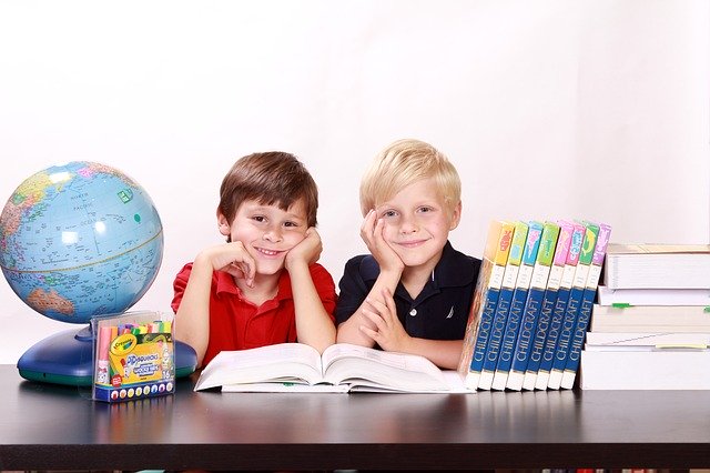 Dos jóvenes sentados en una mesa con libros escolares, un globo terráqueo y lápices de colores, sonriendo a la cámara mientras se preparan para su curso. Su enfoque en el desarrollo de funciones ejecutivas les ayudará a afrontar con éxito los retos futuros.