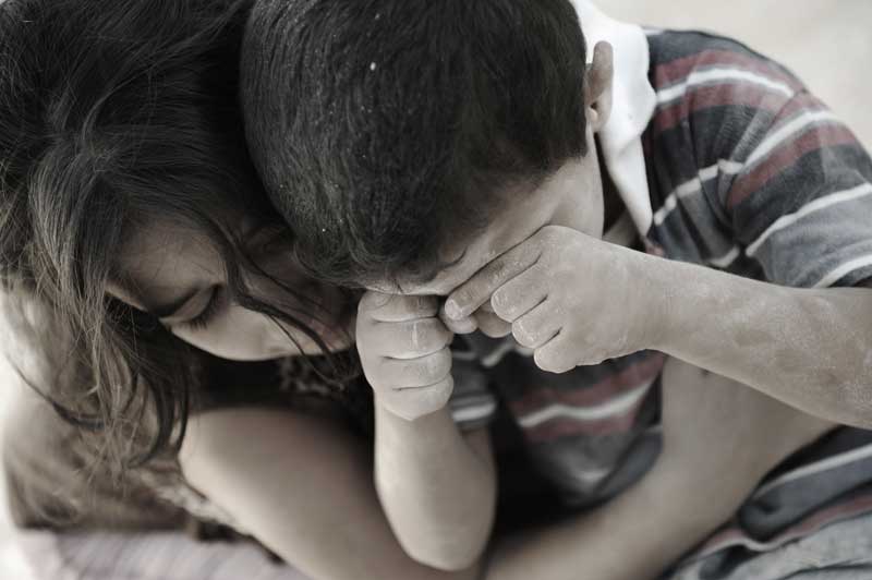 Dos niños están sentados cerca, uno abraza al otro que parece estar secándose los ojos. Ambos visten ropa informal y lucen angustiados, quizás experimentando celos entre hermanos mientras navegan por sus emociones.