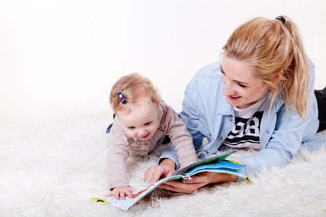 Adulto y bebé tumbados sobre una alfombra blanca, disfrutando de la lectura compartida. El adulto sonríe cálidamente mientras el bebé toca el libro, experimentando los beneficios del vínculo temprano a través de la lectura compartida.