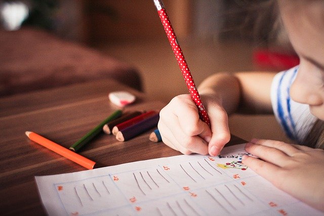 Niño pequeño escribiendo en papel con un lápiz rojo, varios lápices de colores y una goma de borrar sobre la mesa de madera, disfrutando de actividades de apoyo educativo en medio de Vuelta al cole.