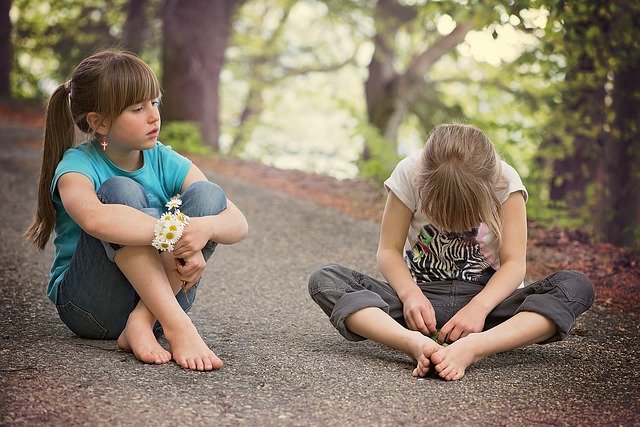 Dos niñas se sientan descalzas en un camino pavimentado rodeado de árboles. Una niña mira hacia un lado mientras la otra, quizás un niño con TEL, se concentra intensamente en un objeto que tiene en la mano.