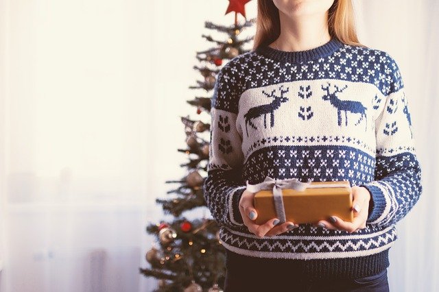 Una persona con un suéter festivo sosteniendo un regalo envuelto, listo para regalar en Navidad, se encuentra frente a un árbol de Navidad bellamente decorado.