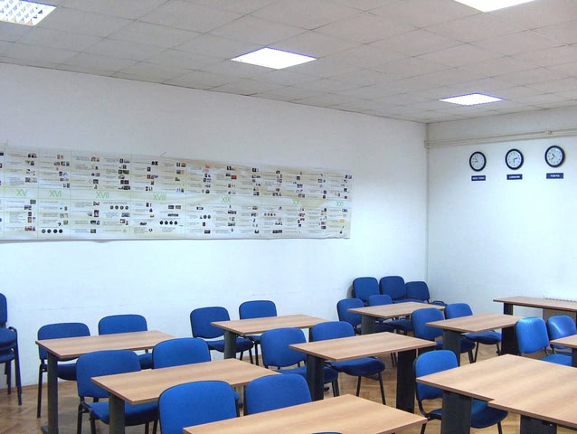 Un salón de clases con sillas azules, escritorios de madera y una cronología de la historia mundial en la pared. Encima de la línea de tiempo hay tres relojes que muestran diferentes zonas horarias, con el objetivo de mejorar la educación y reducir el fracaso escolar en España.