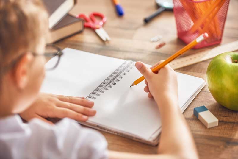 Una persona con gafas escribe en un cuaderno abierto sobre un escritorio de madera con artículos de papelería dispersos, entre ellos una manzana, una goma de borrar, una regla, libros y un portalápices rojo. La escena evoca el minucioso proceso de adaptación seguido en un centro de educación infantil.