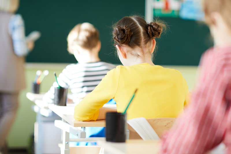 Los niños están sentados en pupitres en un salón de clases, frente a una pizarra, listos para el comienzo del año escolar.