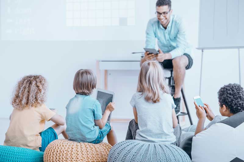 Un profesor sentado en un taburete frente al aula, sonriendo y sosteniendo una tableta. Cuatro estudiantes se sientan sobre cojines y cada uno utiliza una tableta o un teléfono inteligente. Esta escena resume perfectamente el entorno de aprendizaje moderno, un factor importante cuando buscas elegir una escuela para tu hijo.