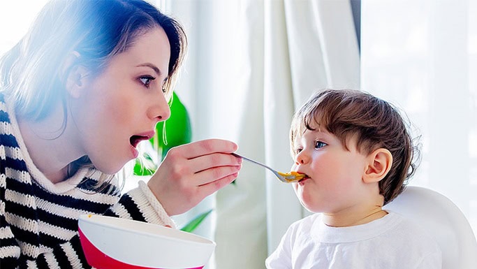 Un adulto alimenta a un niño pequeño con una cuchara en una trona, gestionando cuidadosamente el proceso debido a la DISFAGIA NEONATAL. El adulto sostiene un cuenco y ambos se concentran en garantizar que el niño trague con seguridad.