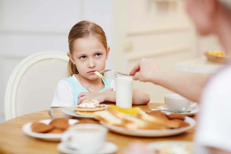 Una niña sentada en una mesa de comedor con platos de comida, luciendo disgustada. Se ve la mano de un adulto sosteniendo un tenedor cerca de ella, lo que sugiere que el impacto del confinamiento puede estar contribuyendo a posibles trastornos alimentarios.