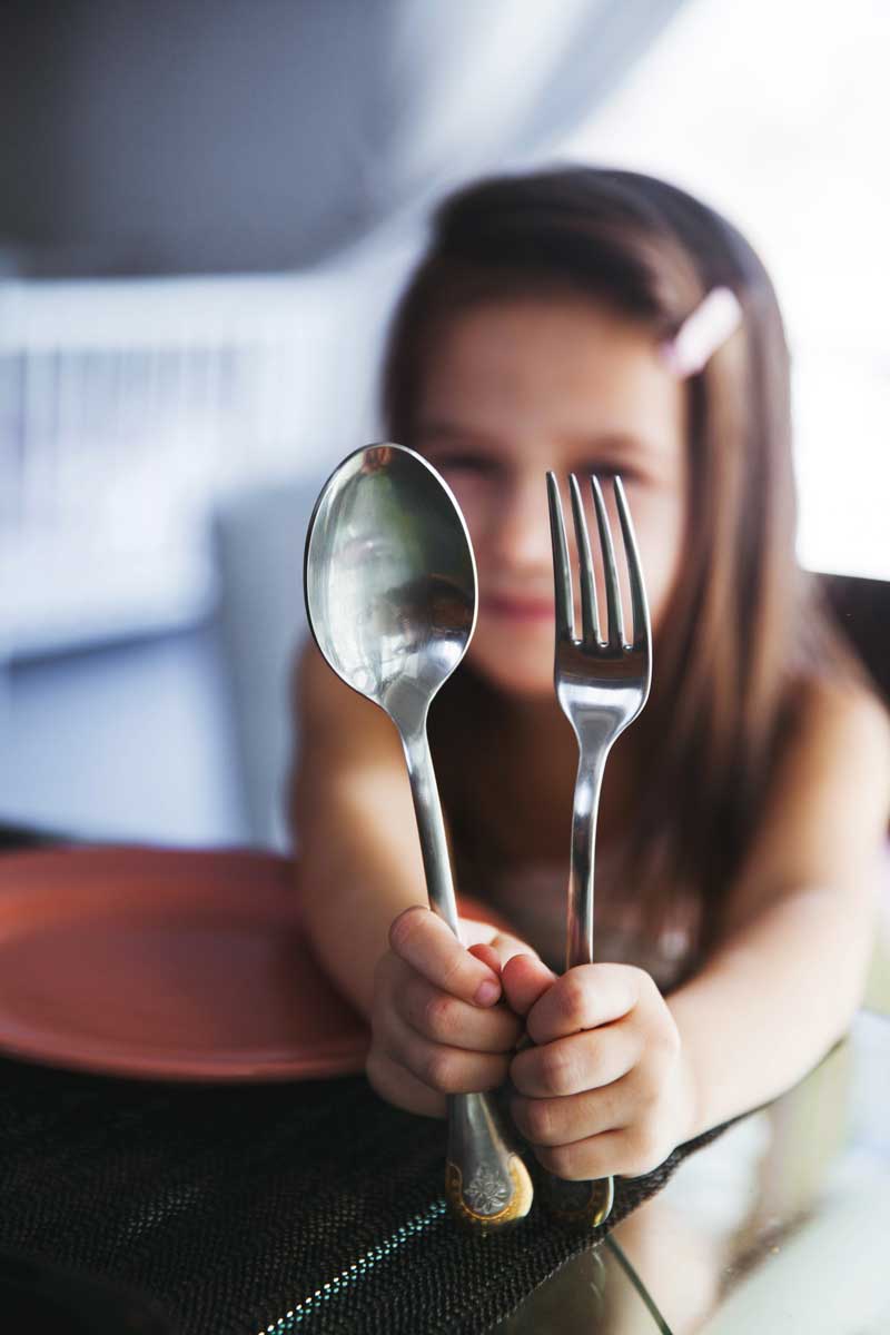 Una niña en su infancia sostiene cubiertos (una cuchara y un tenedor) frente a su cara, con platos borrosos y una mesa al fondo.