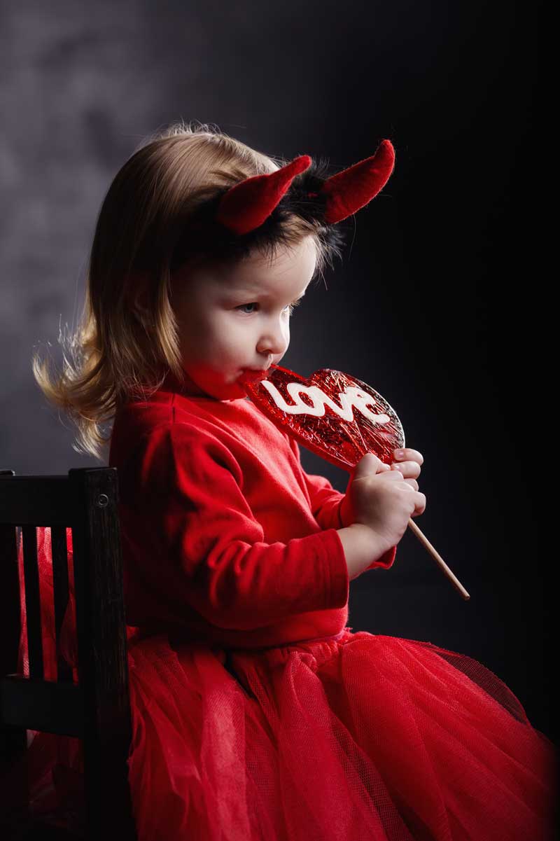 Un niño pequeño con un traje rojo y cuernos de diablo en una diadema sostiene una paleta "LOVE" mientras está sentado en una silla sobre un fondo oscuro, simbolizando el equilibrio lúdico entre la picardía y el desarrollo moral.