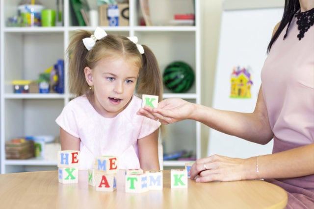 Una niña con coletas interactúa con un adulto, jugando con bloques de letras, en una mesa en un preescolar o guardería, enfocándose en técnicas de logopedia para abordar posibles secuelas del desarrollo del habla.