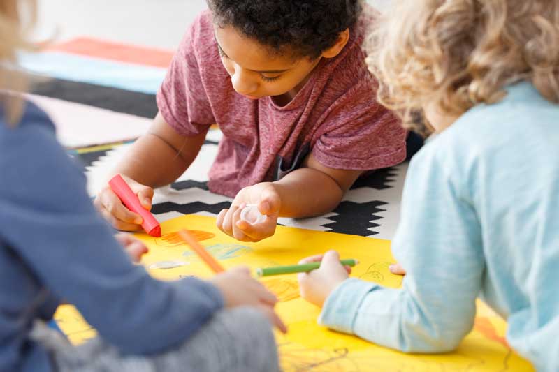 Tres niños están creando dibujos juntos en un papel amarillo grande usando marcadores de colores. Están sentados en el suelo con una alfombra estampada en blanco y negro debajo, dando vida a sus imaginativas interpretaciones.