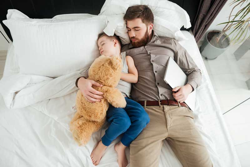 Un hombre y un niño duermen una siesta en una cama. El niño abraza un osito de peluche y el hombre, que a menudo tiene problemas de sueño, sostiene una tableta con una mano.