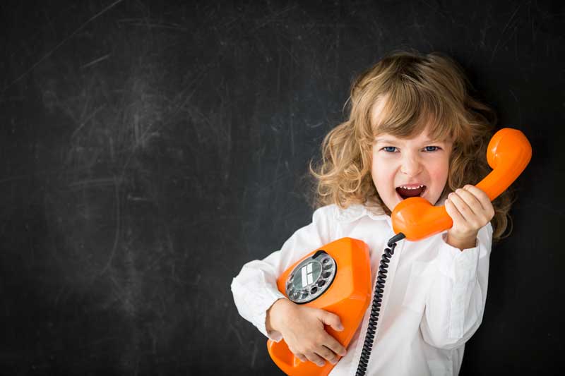 Una niña de pelo largo y camisa blanca sostiene un teléfono de disco naranja sobre un fondo oscuro y su expresión oscila entre la emoción y el enfado.