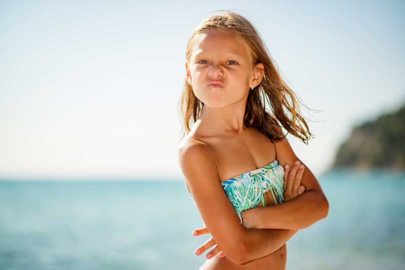 Una niña en traje de baño está parada en una playa con los brazos cruzados, haciendo una cara juguetona con los labios fruncidos. Al fondo se ven borrosos el océano y una costa lejana.