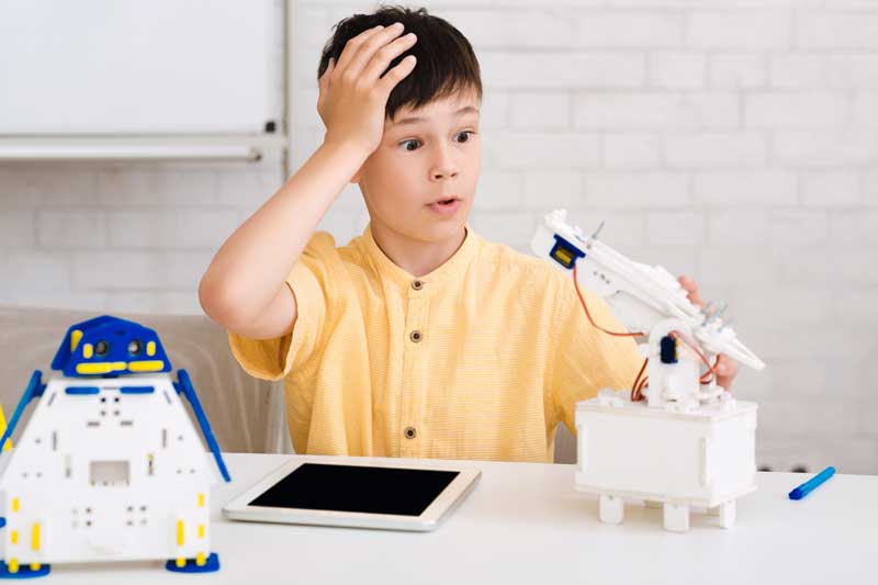 Un niño con una camiseta amarilla parece sorprendido mientras interactúa con un brazo robótico, una tableta y otro pequeño robot en la mesa frente a él, como si reconociera el potencial de errores cometer.