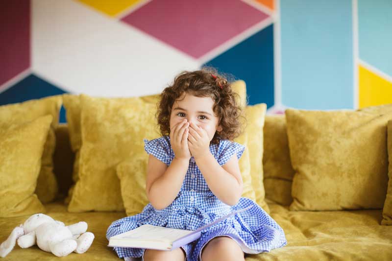 Una niña pequeña con un vestido azul sentada en un sofá amarillo, tapándose la boca con ambas manos, mostrando características de ECOLALIA. A su lado hay un peluche y un libro abierto. Al fondo hay una colorida pared geométrica.