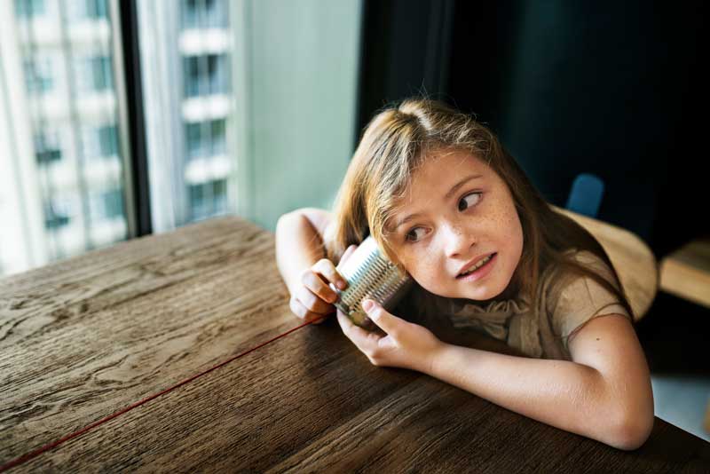 Una niña escucha a través de un teléfono de lata mientras está sentada en una mesa de madera cerca de una ventana, practicando su comprensión oral.