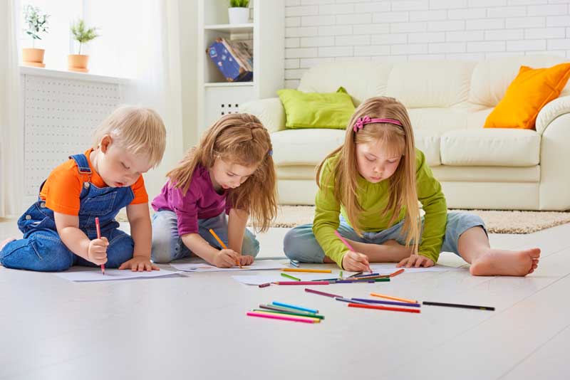 Tres hijos están tirados en el suelo, dibujando con lápices de colores sobre papel. A su alrededor hay varios lápices de colores, creando una escena vibrante. Al fondo, un sofá con cojines de colores ofrece un acogedor telón de fondo para su entrenamiento creativo.