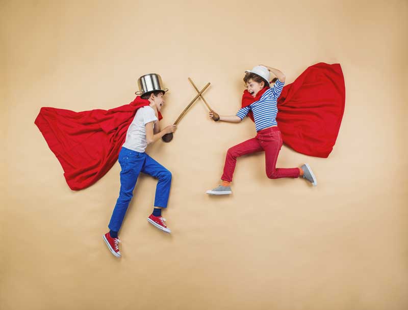 Dos niños con capas y gorros rojos simulan luchar con palos de madera tumbados sobre una superficie beige, fomentando la estimulación y el desarrollo en sus hijos.